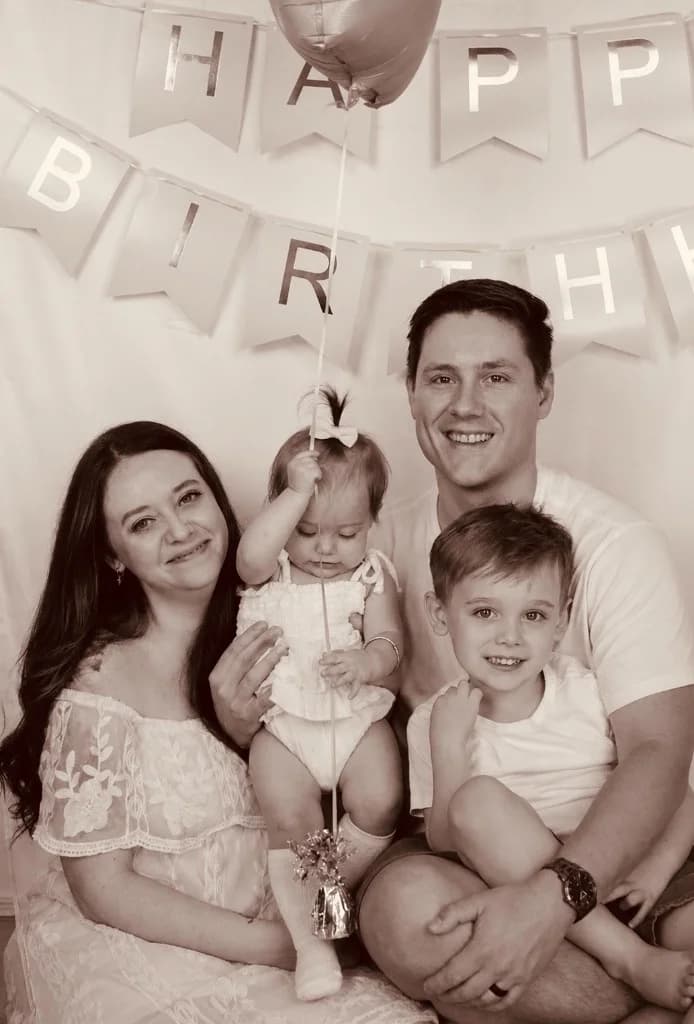 Chelsea Dietz with her family — husband, daughter holding a balloon, and son — in front of a 'Happy Birthday' banner.